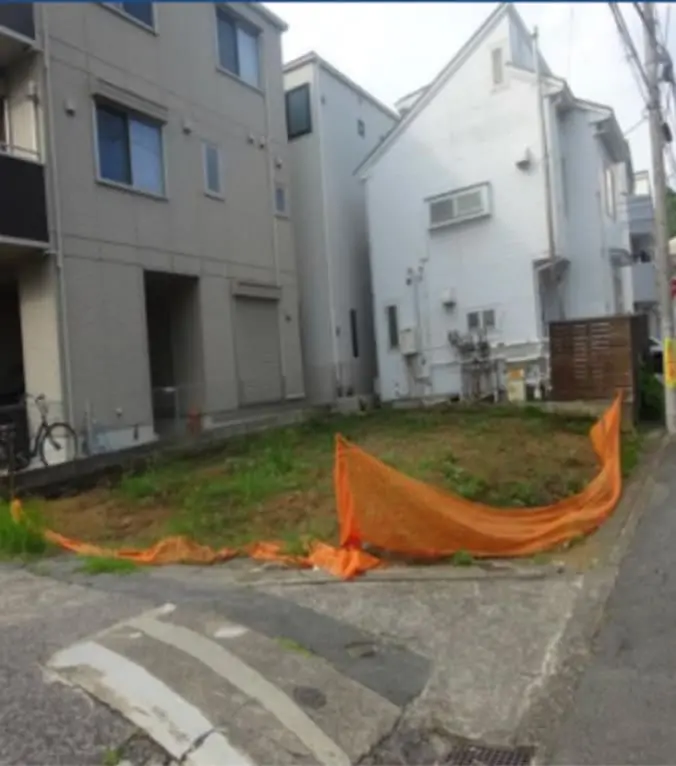 住宅地にある更地の写真で、周囲には戸建てやアパートが隣接し、工事用のネットが張られている
                                                